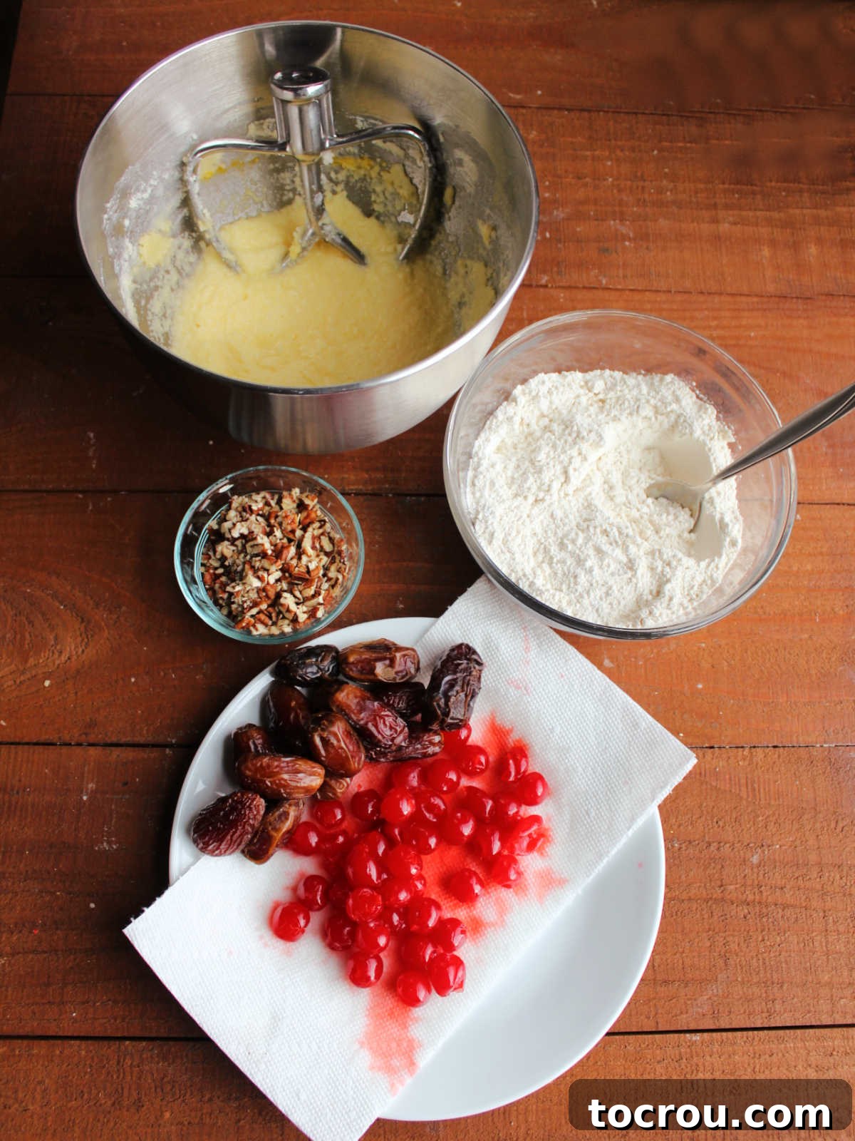 Making the cherry winks dough with a mixer bowl with the wet ingredients, a mixing bowl with the dry ingredients, a bowl of chopped pecans, and a plate with drained maraschino cherries and dates.