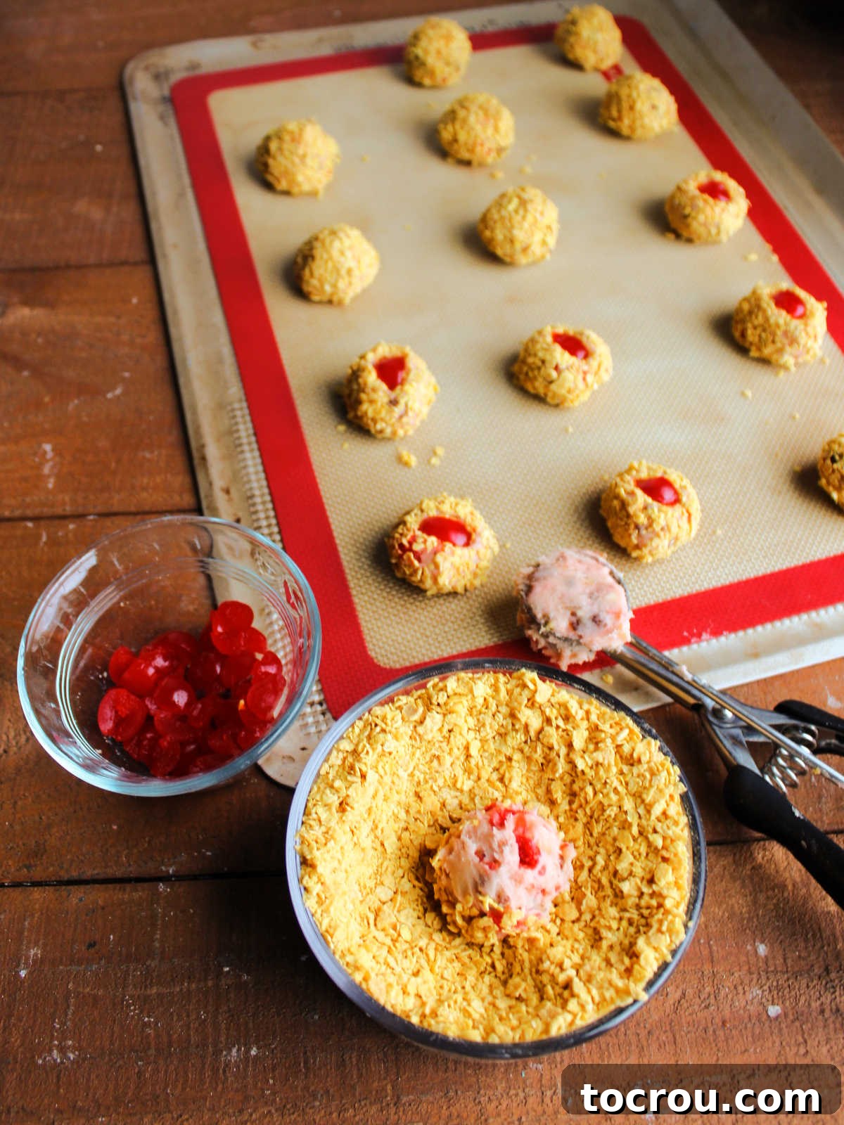 Scooping dough into a bowl of crushed cornflakes to coat the exterior with a bowl of slivered maraschino cherries nearby and a baking tray with finished cookies in the background, ready to go in the oven. 