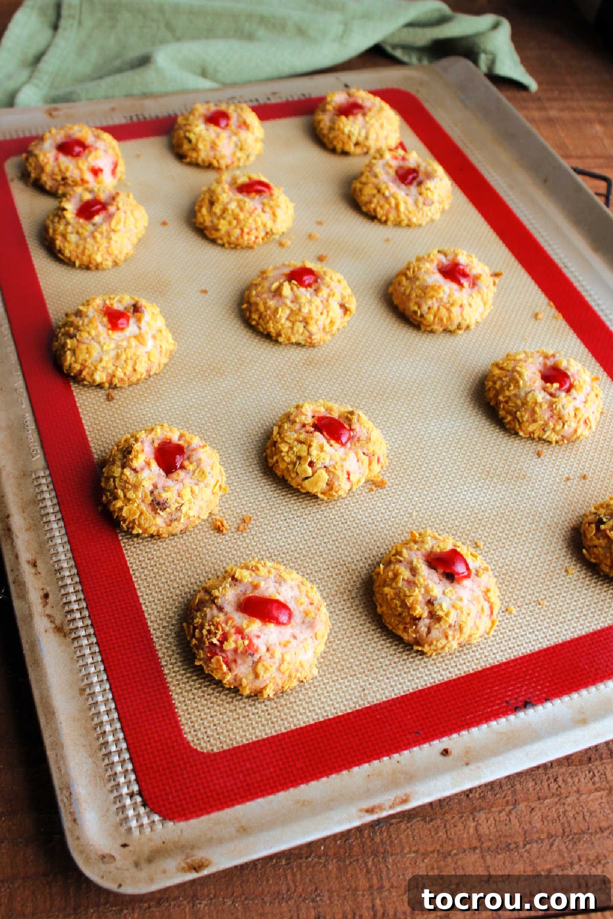 Cookie sheet lined with a silicone mat topped with freshly baked cherry wink cookies expanded slightly so the pink dough shows through the cornflake exterior. 