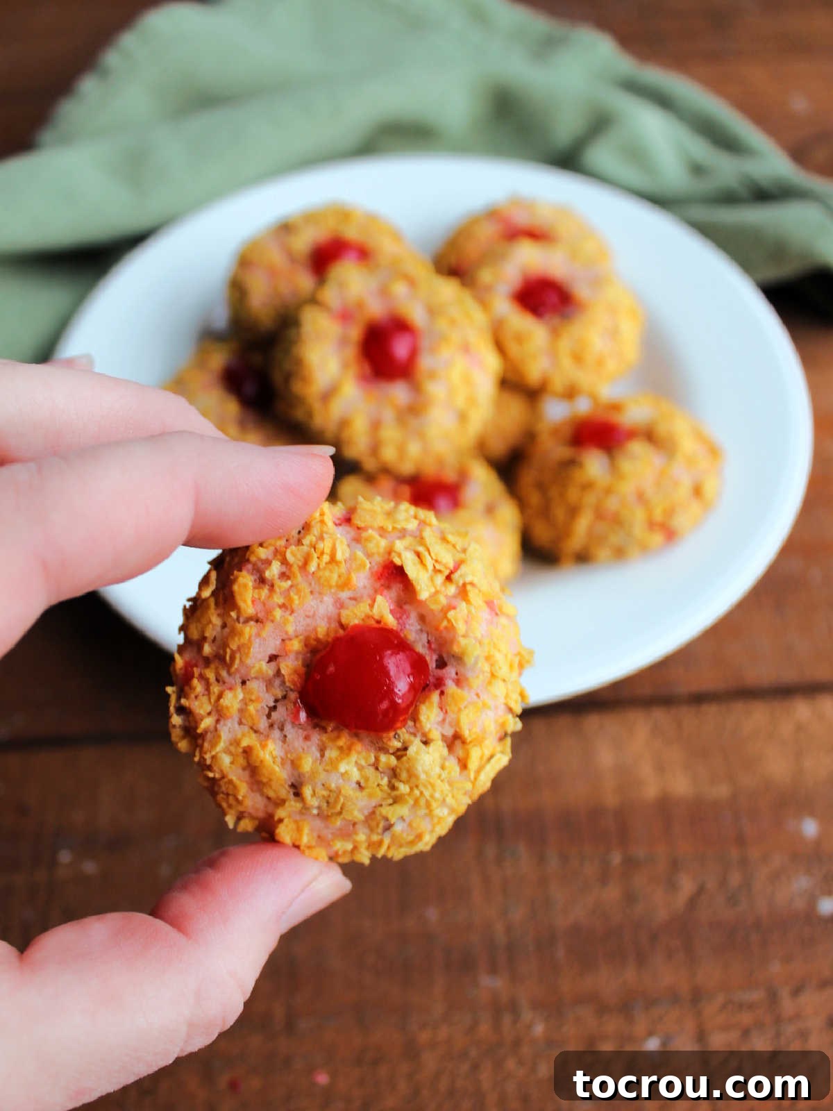 Hand holding a cherry wink cookie with a plate of more cookies in the background. 