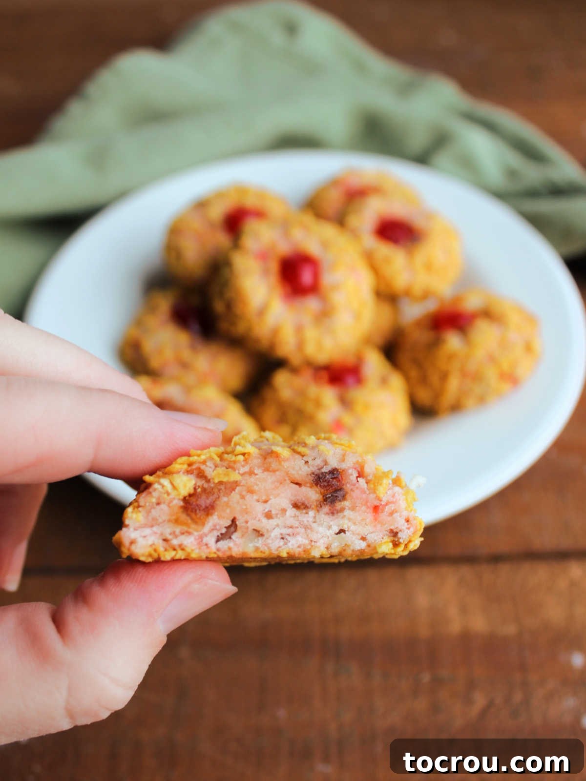 Hand holding half of a cherry wink cookie showing the bits of cherry, dates, and pecans inside the light pink cookie and the cornflake bits on the outside. 