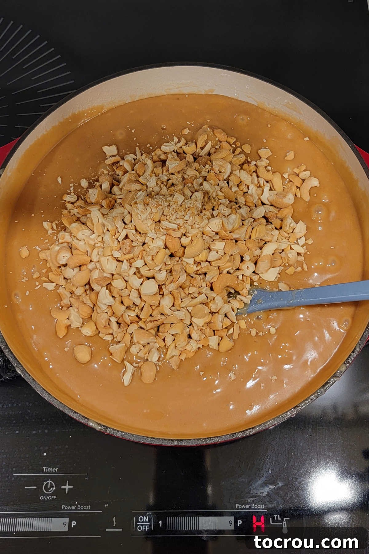 A cast iron pan on the stovetop, filled with warm, bubbling, homemade gooey caramel and freshly chopped cashews, ready to be stirred.