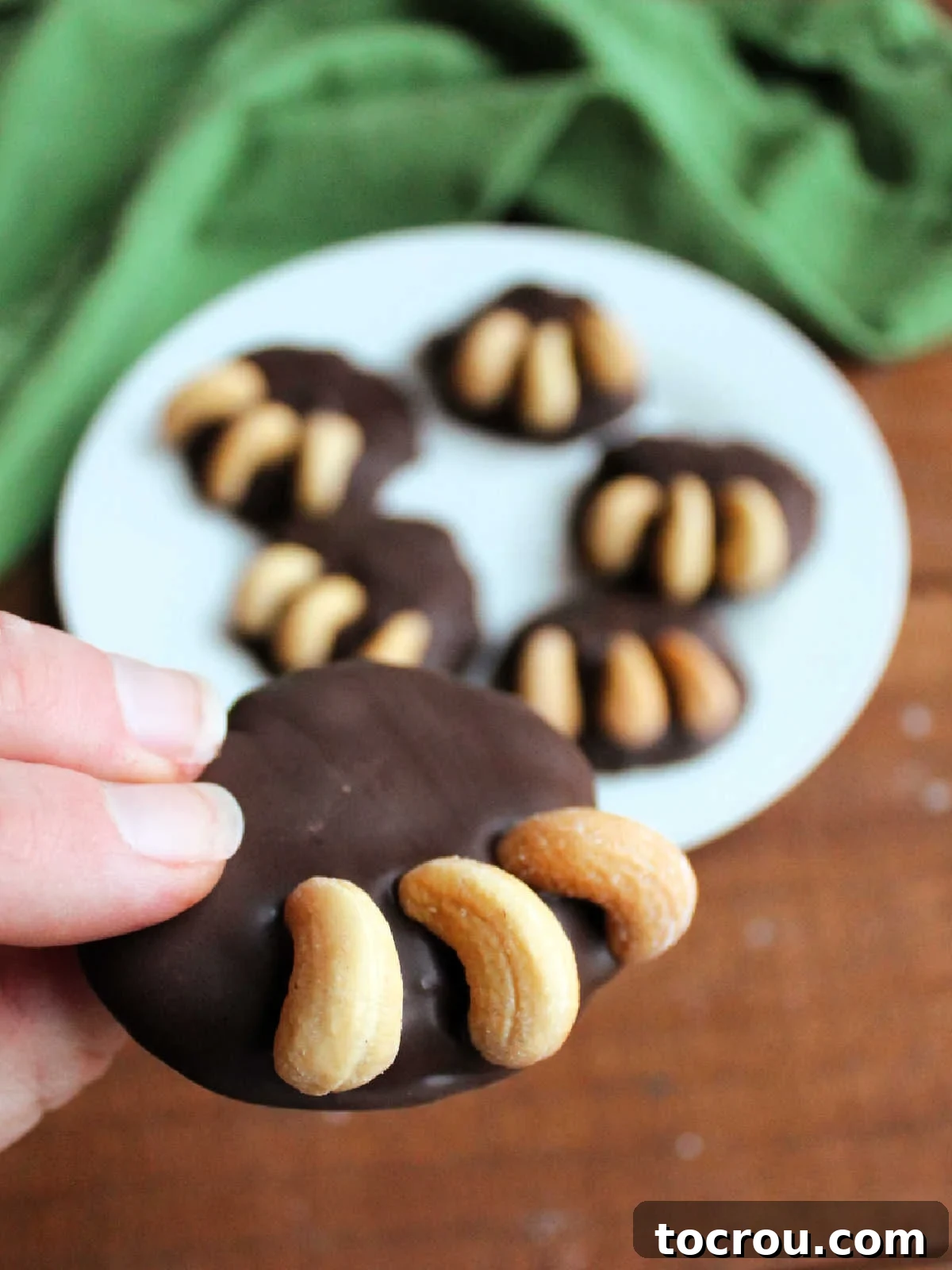 A hand gently holds a single, perfectly formed bear paw candy, showcasing its rich chocolate-dipped caramel and cashew center, beautifully adorned with three additional cashews arranged to resemble a bear claw.