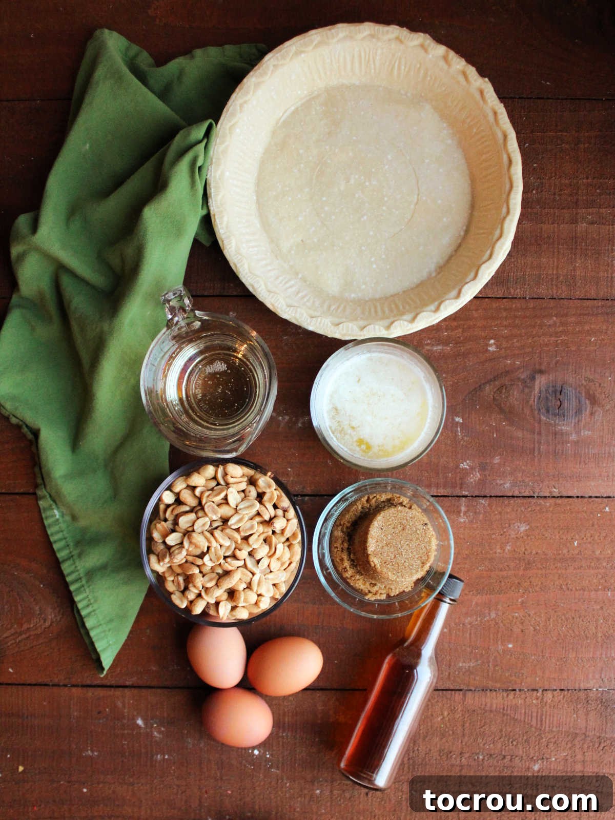 Ingredients for old fashioned peanut pie: pie crust, corn syrup, melted butter, brown sugar, vanilla, eggs, and roasted peanuts arranged on a wooden surface.