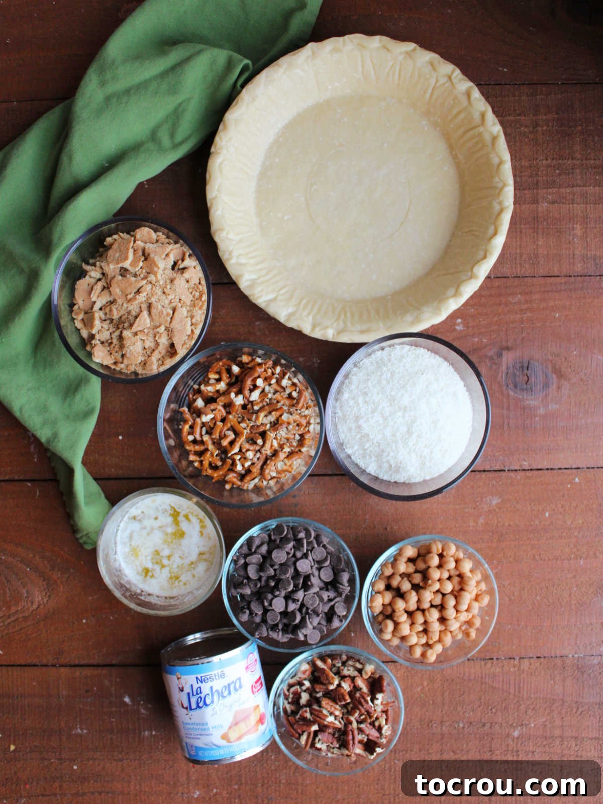 Magic Cookie Bar Pie Ingredients All the essential ingredients for Magic Cookie Bar Pie laid out: a store-bought pie crust, caramel bits, vibrant chocolate chips, crunchy pecans, crushed graham cracker pieces, salty pretzel pieces, melted butter, and a can of sweetened condensed milk, ready for assembly.