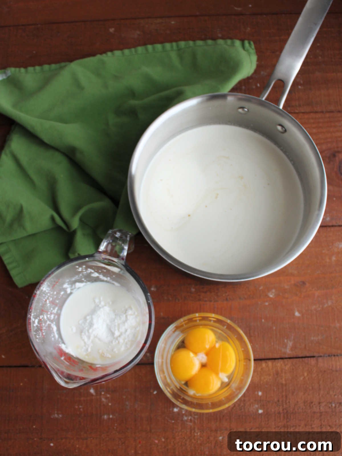 Making maple pie filling with saucepan with maple syrup, brown sugar, milk and cream next to measuring cup with milk and cornstarch and a small bowl of egg yolks. 