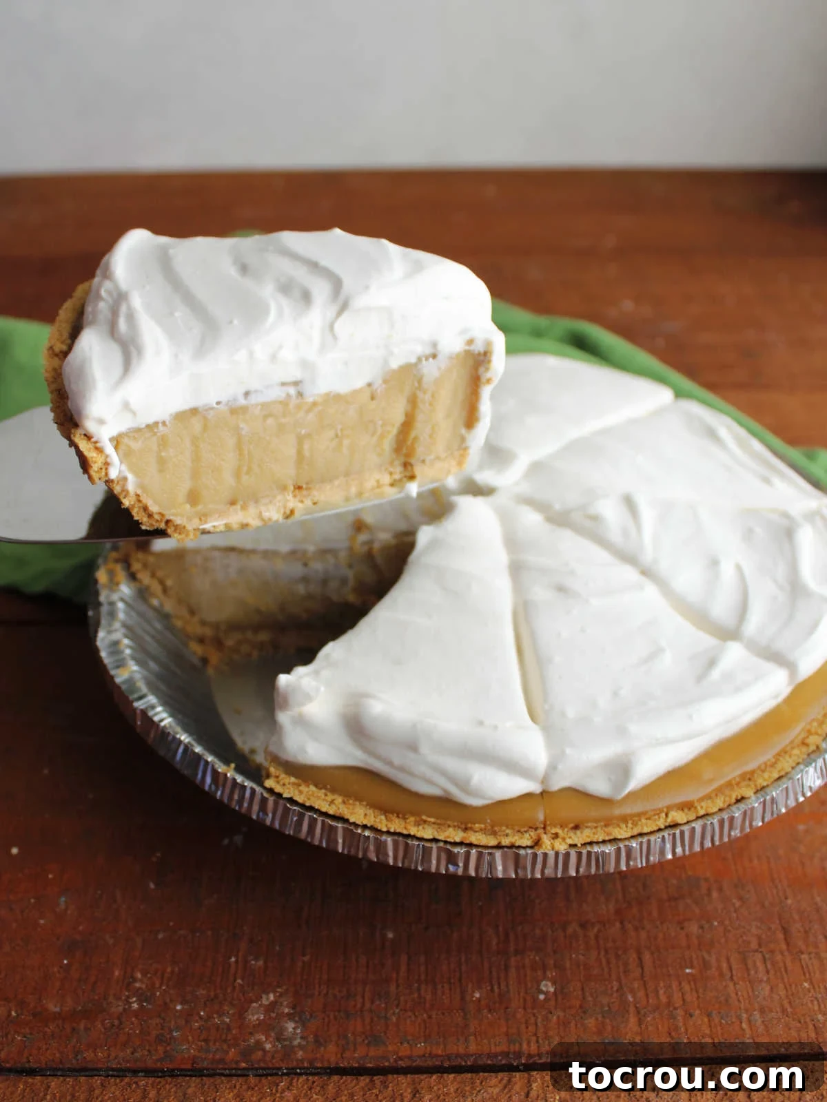 Pie server lifting first slice of whipped cream topped maple cream pie out of the pie plate showing the thickened custard filling. 