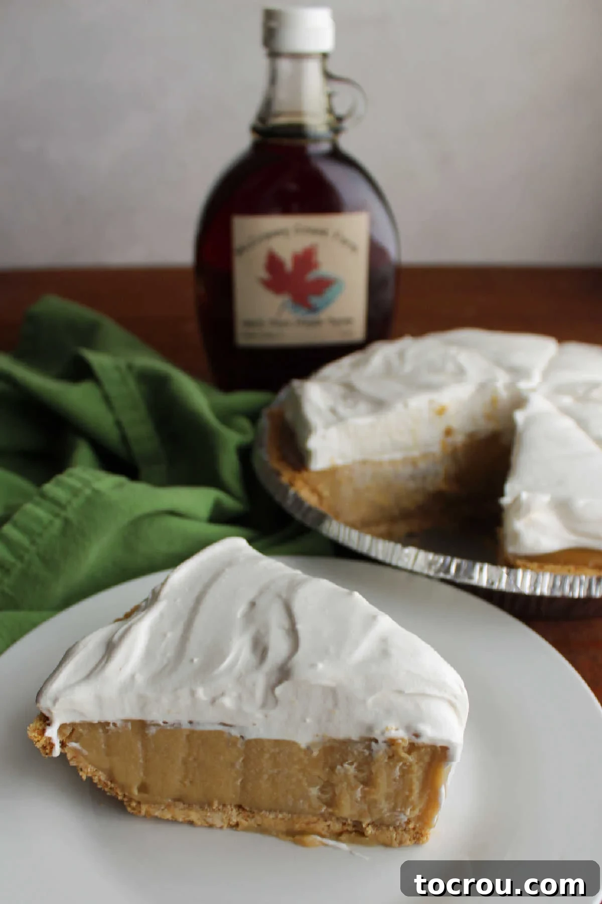Slice of homemade maple cream pie on plate with remaining pie and a bottle of pure maple syrup in the background. 