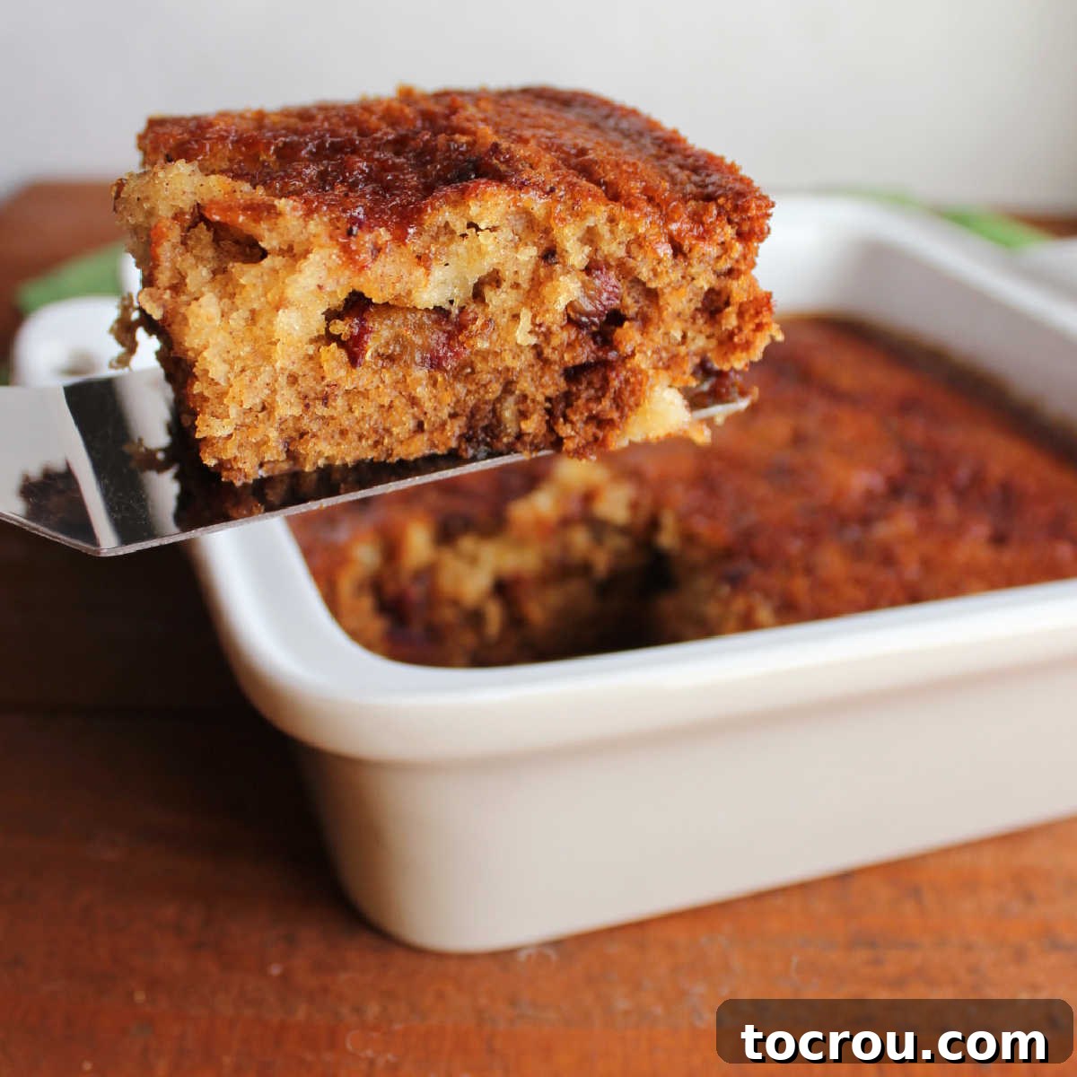Spatula lifting piece of date pudding out of square baking dish showing bits of date in sticky cake.