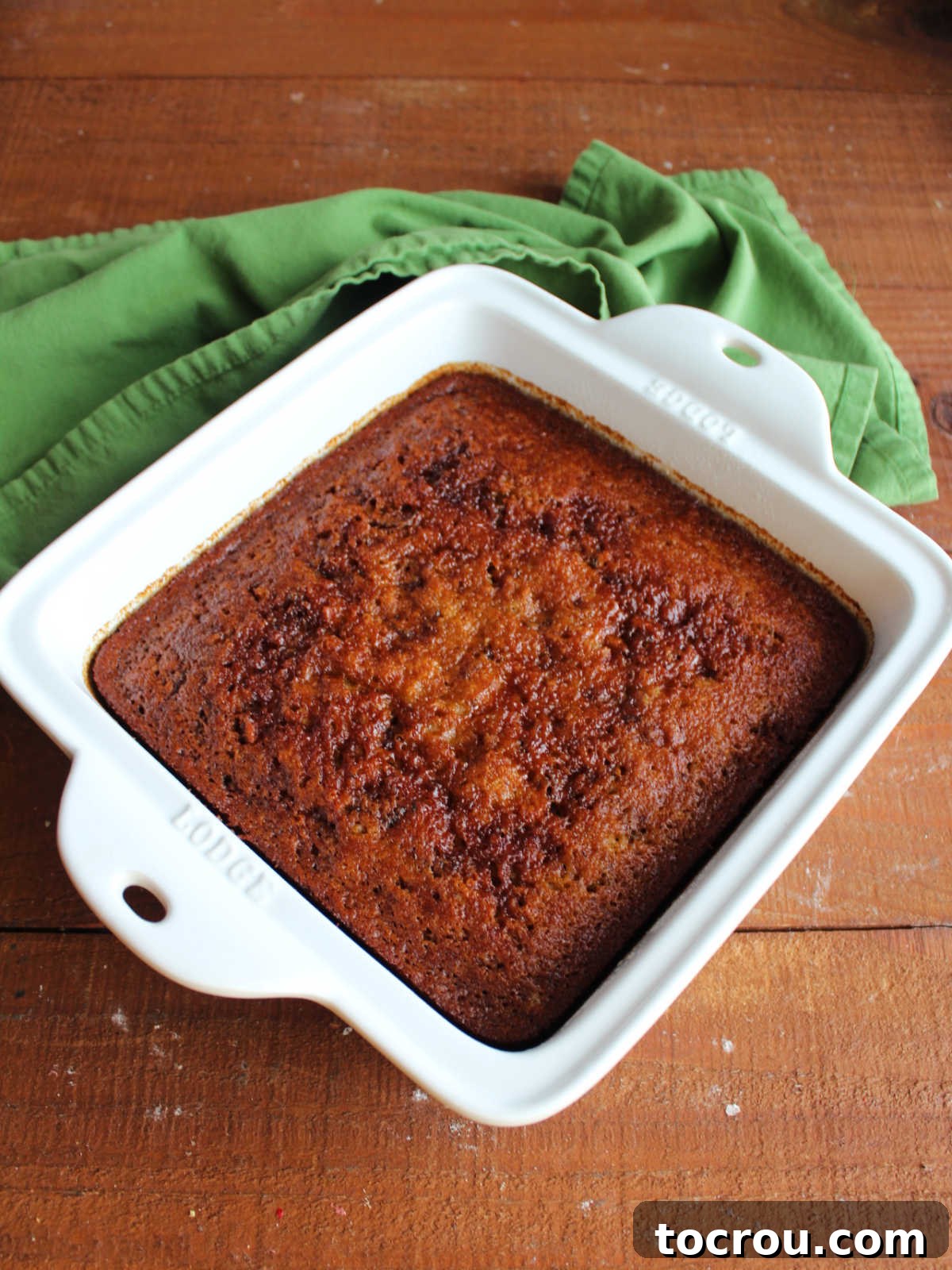 Baked date pudding in square baking dish fresh from the oven showing dark brown top and gooey texture. 