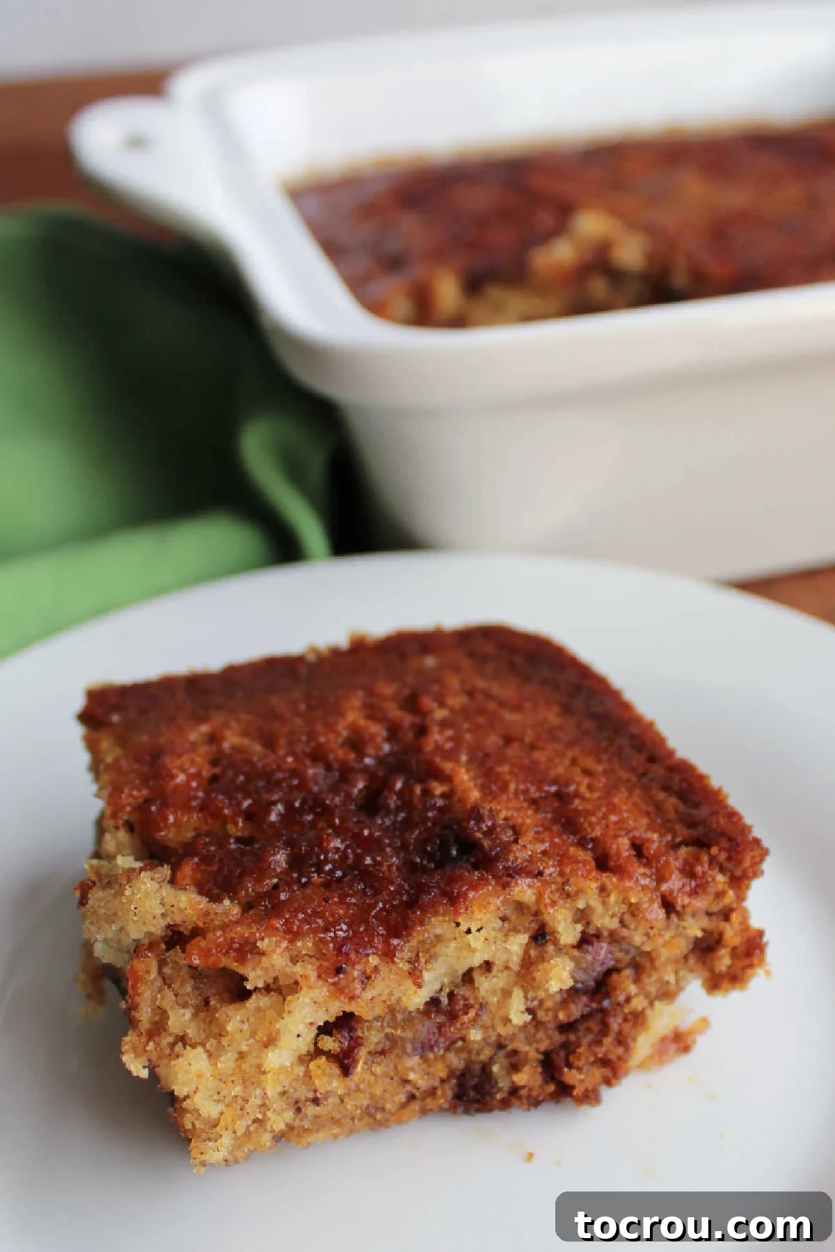 Piece of date pudding with a cake like texture and bits of dates on a plate with pan of remaining pudding in the background. 