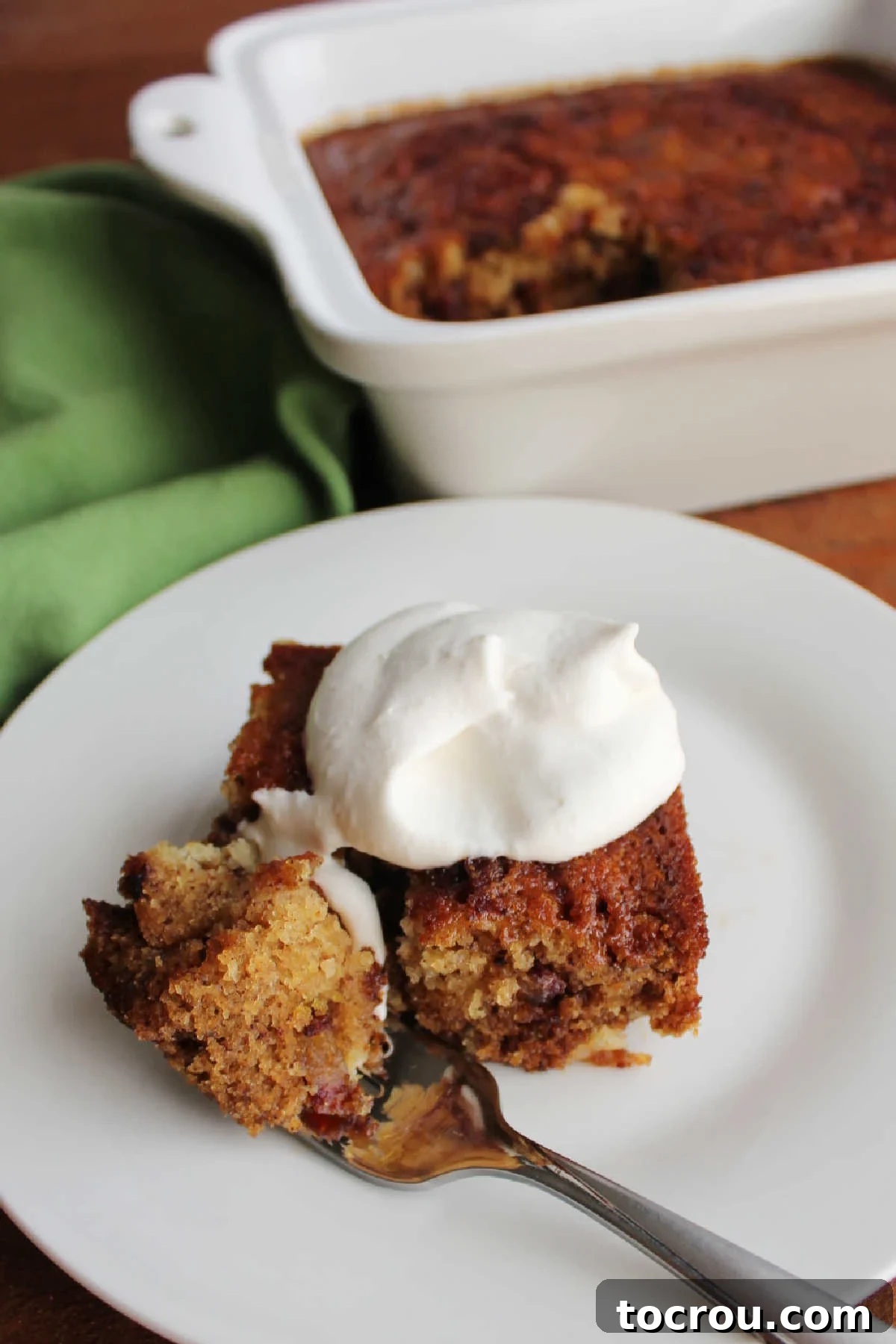 Bite of sticky date pudding on fork next to piece of date pudding topped with maple whipped cream, ready to eat. 
