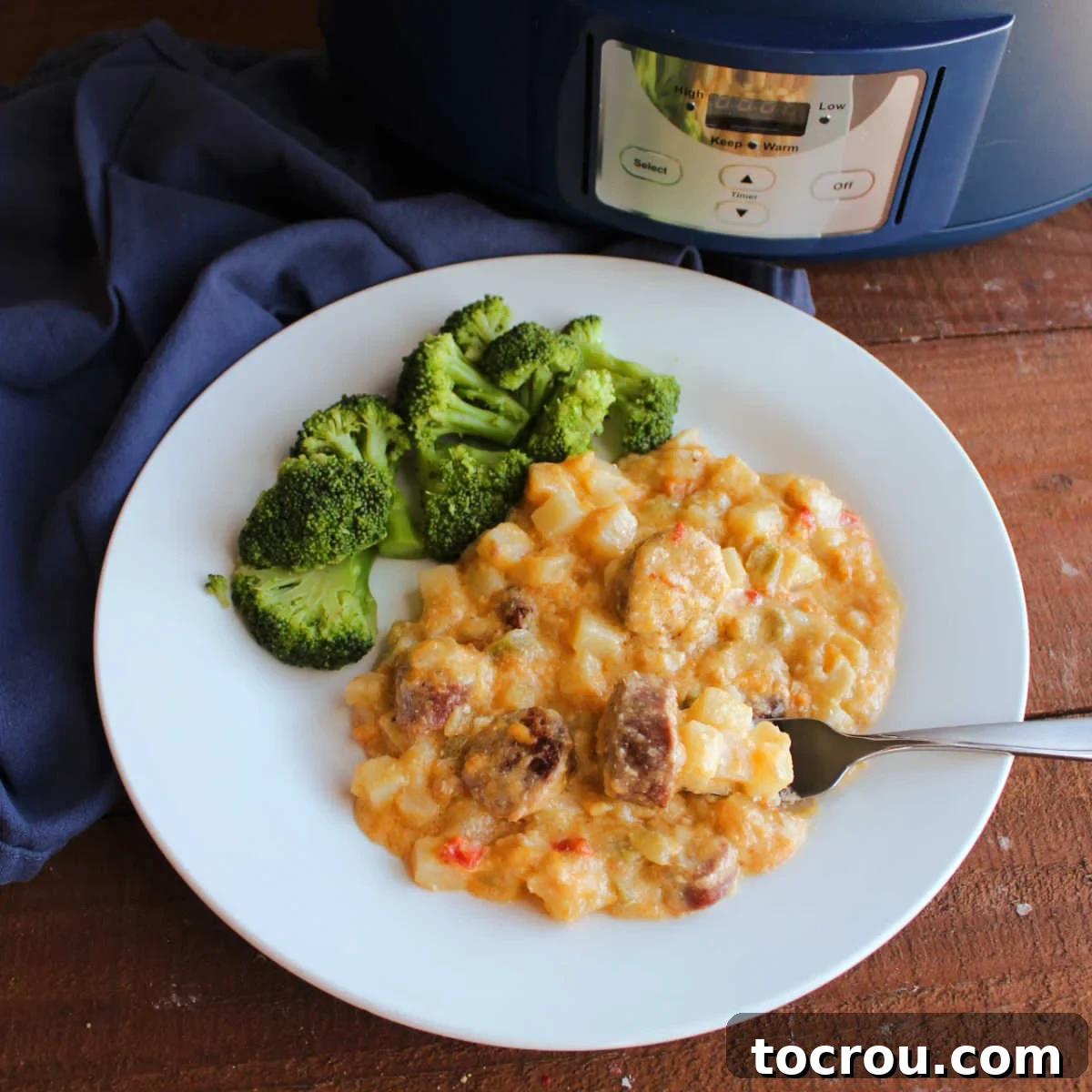 A serving of creamy cheesy potatoes with chunks of kielbasa, garnished with fresh parsley, alongside steamed broccoli on a dinner plate. A slow cooker is blurred in the background, hinting at the meal's effortless preparation.