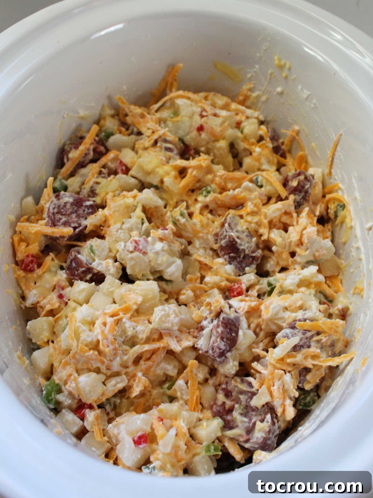 The ingredients for crockpot cheesy potatoes and kielbasa, including frozen hash browns, sliced smoked sausage, cream of chicken soup, sour cream, and shredded cheese, have been gently stirred together inside the crockpot insert, ready for cooking.