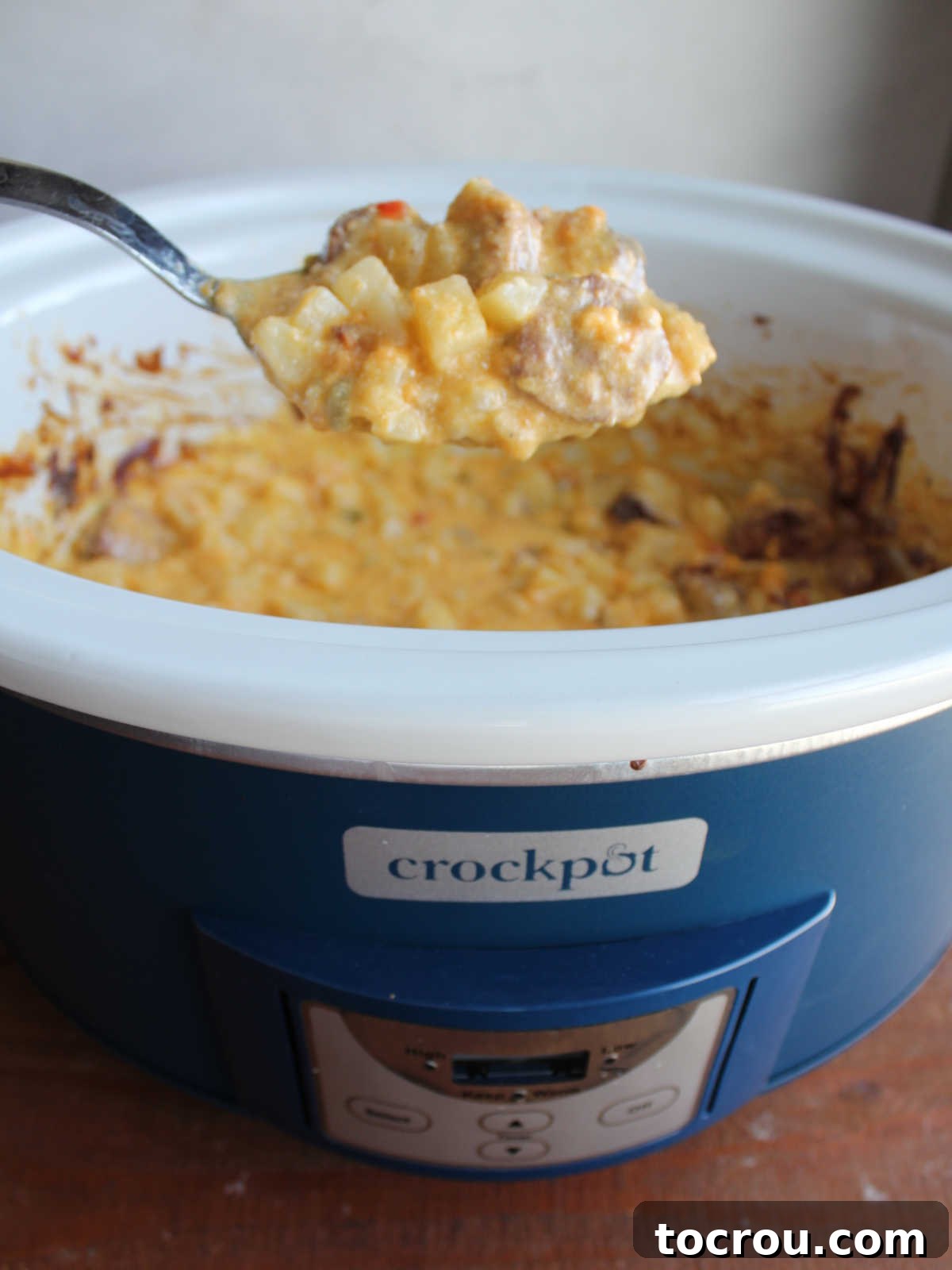 A serving spoon lifting a generous scoop of creamy cheesy potatoes and smoked sausage from the crockpot, showcasing the thick, luscious sauce that developed during slow cooking.
