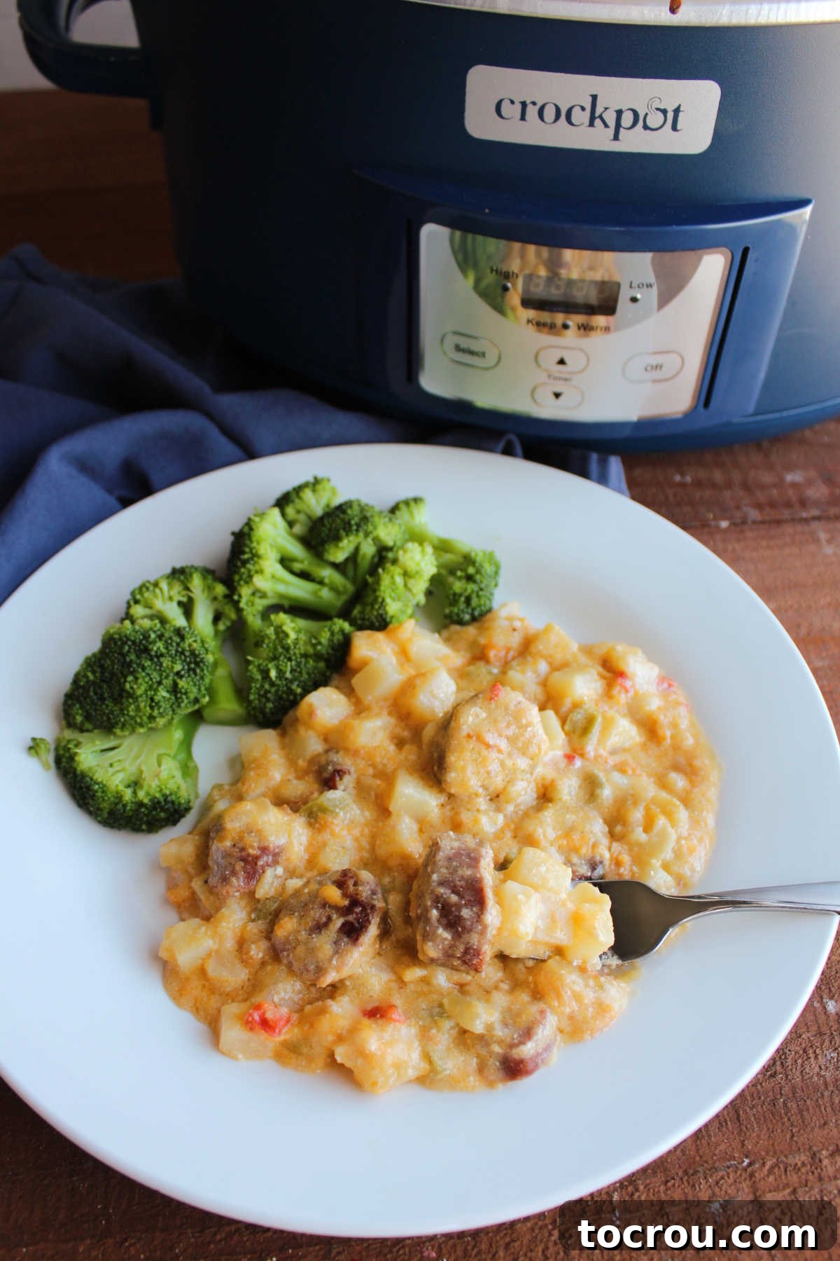 A cozy dinner plate showcasing a generous serving of cheesy potatoes and smoked sausage, accompanied by a vibrant side of steamed broccoli. The slow cooker in which the meal was cooked sits invitingly in the background, ready for seconds.