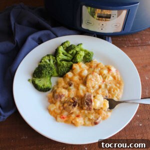 Plate of cheesy potatoes with chunks of kielbasa next to steamed broccoli on dinner plate with crockpot in the background.