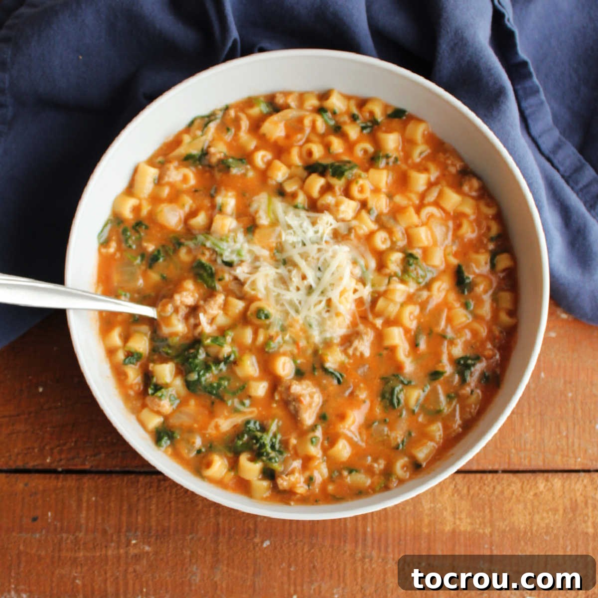 Bowl of soup with creamy tomato base, bits of Italian sausage, ditalini pasta, and spinach topped with Parmesan cheese.