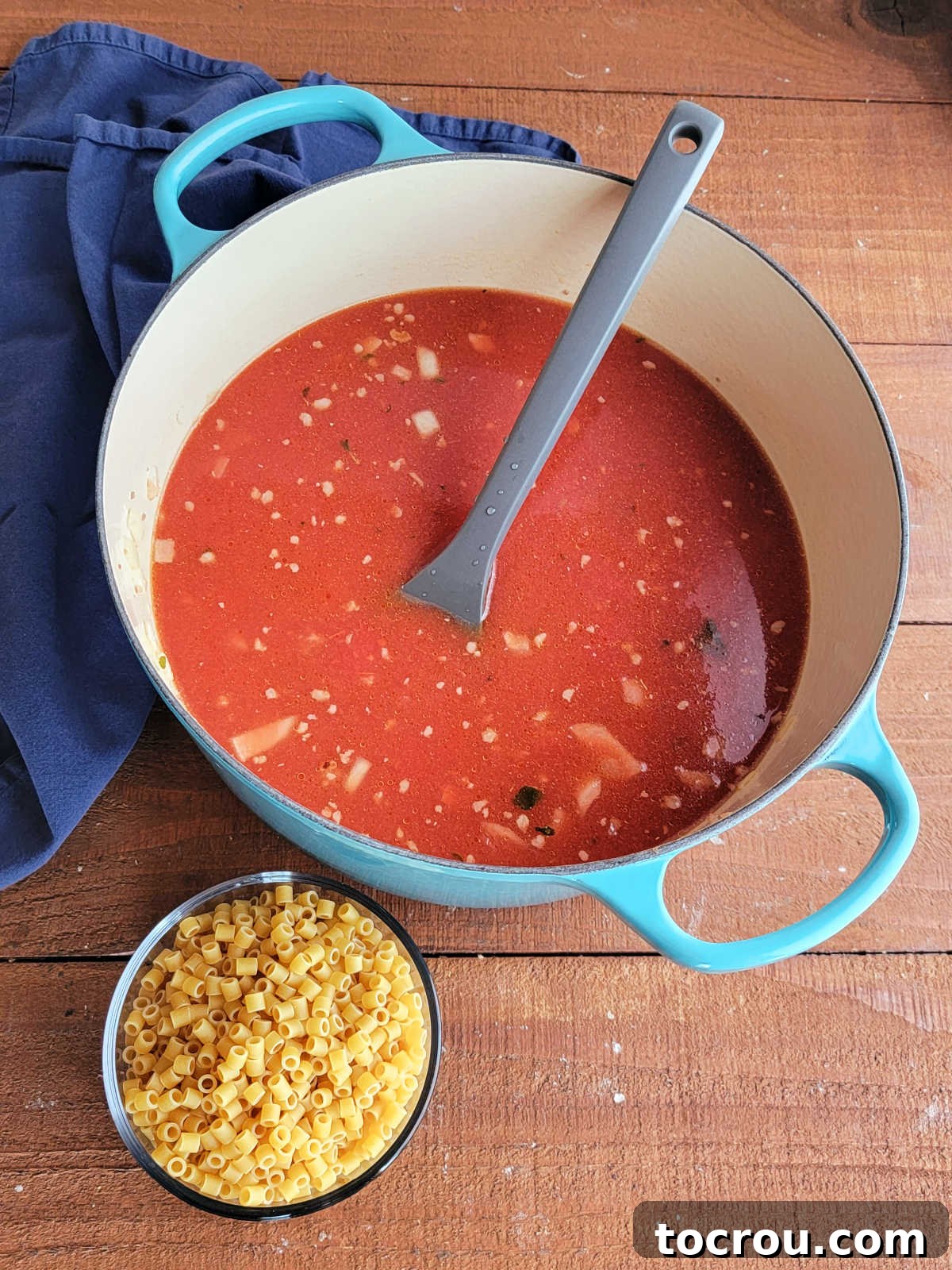 Dutch oven with tomato broth and sausage inside next to a bowl of raw ditalini pasta, ready to go in the pot to cook. 