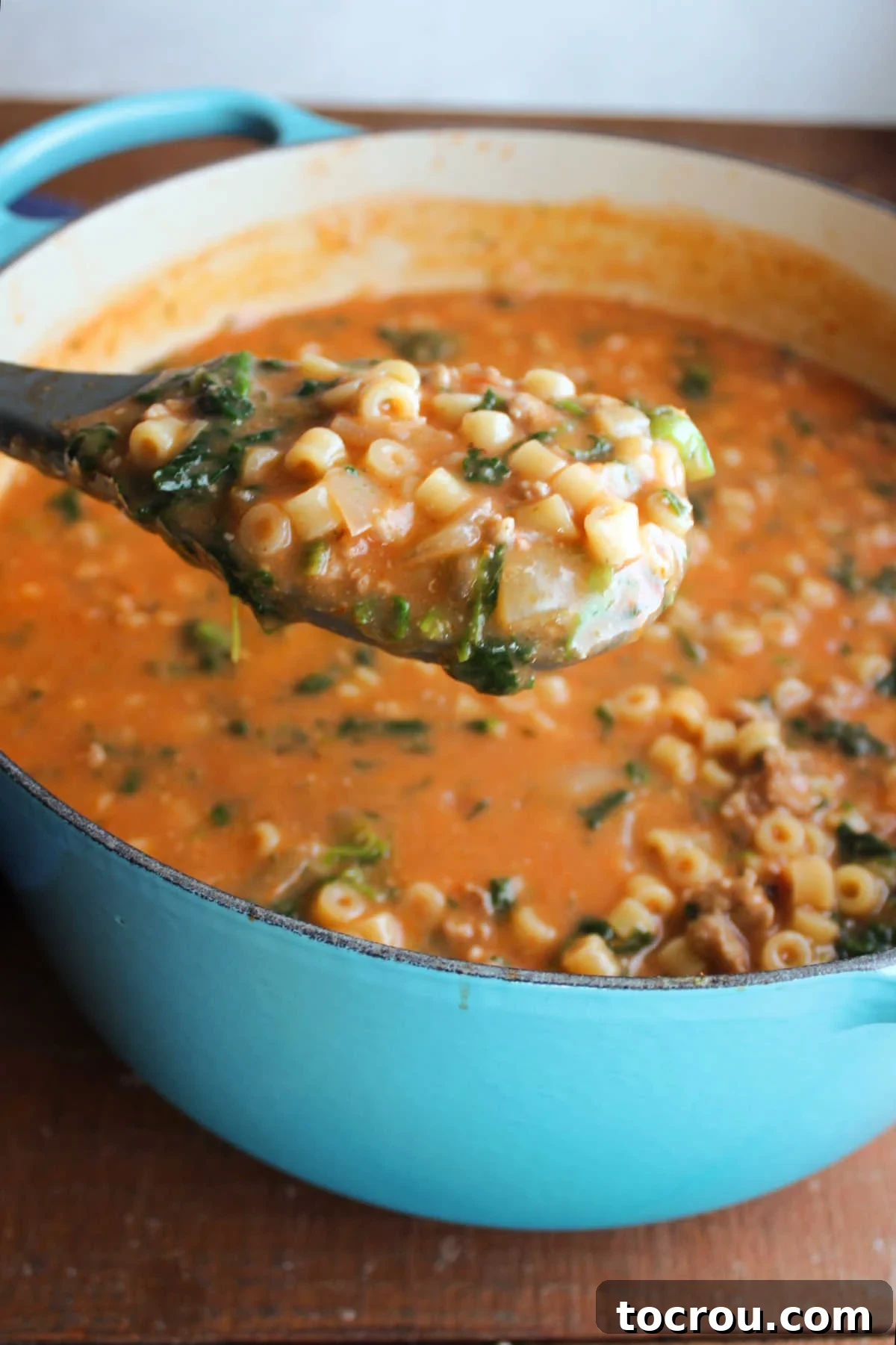 Serving spoon lifting a scoop of Italian sausage and ditalini soup out of Dutch oven with more soup inside. 