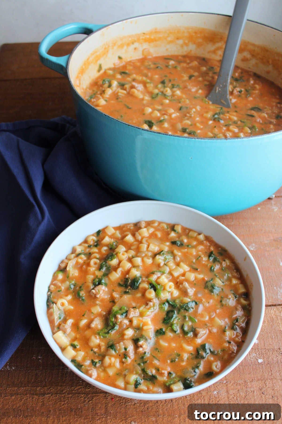 Bowl of Italian sausage soup with ditalini pasta and spinach in creamy tomato broth next to the dutch oven it was cooked in.