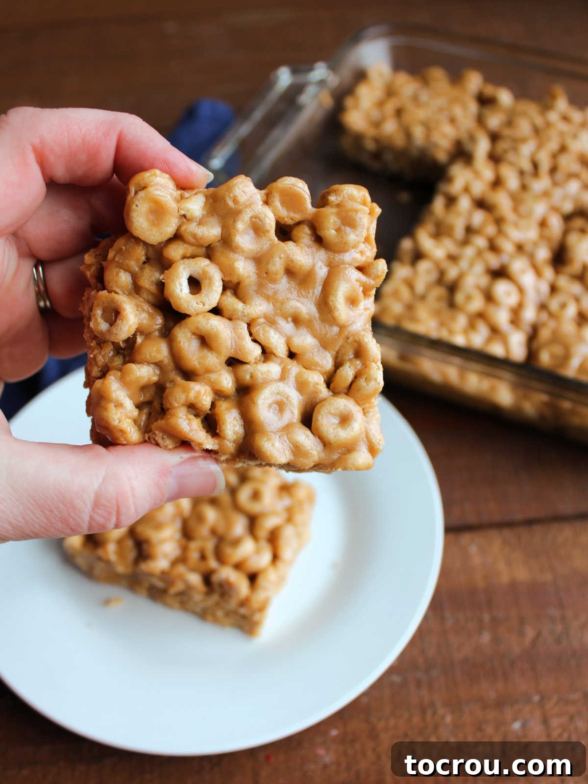 Close-up of Cheerio Bar A close-up shot of a hand holding a single square of a no-bake peanut butter Cheerio bar, showcasing the individual Cheerios bound together by the thick, chewy peanut butter mixture.