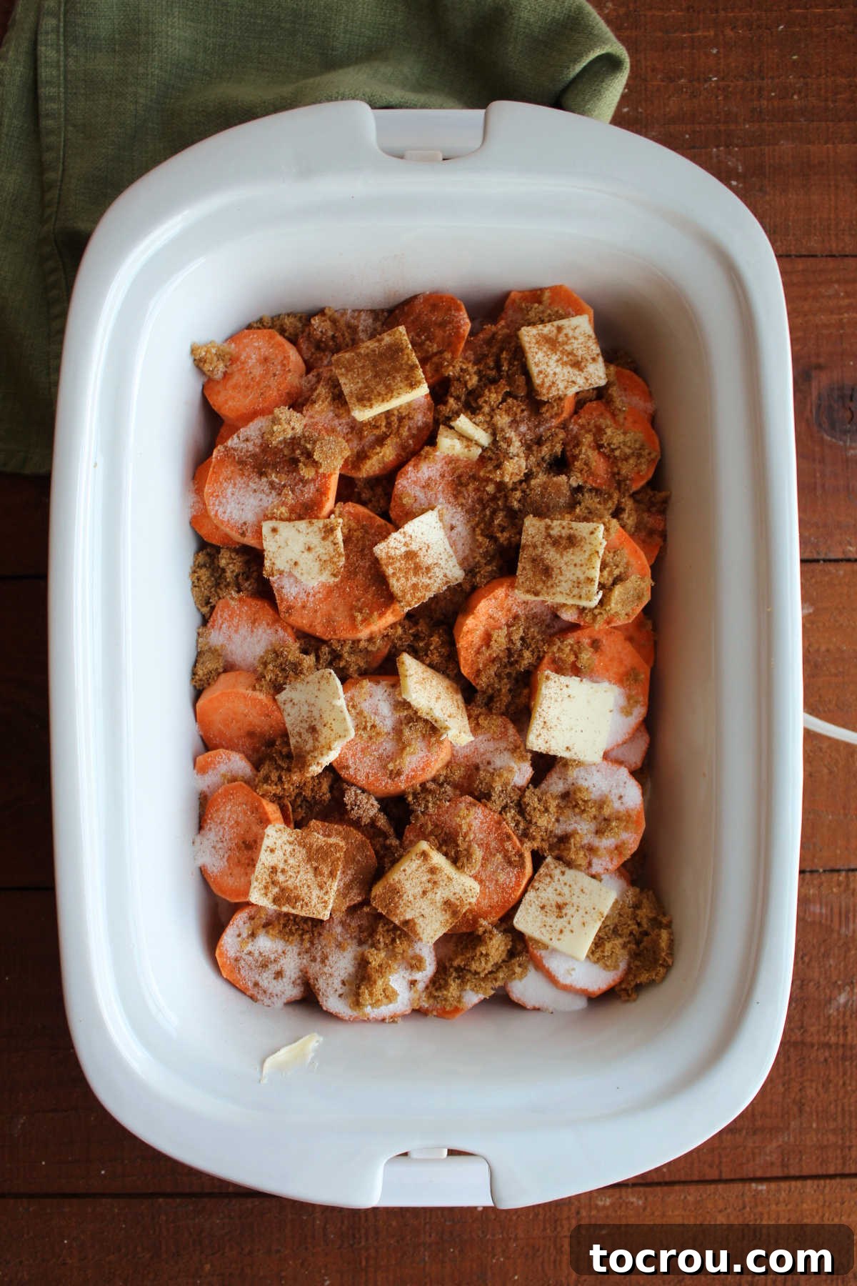 Crockpot filled with slices of fresh sweet potatoes topped with sugar, brown sugar, butter, cinnamon, and nutmeg, ready to cook. 