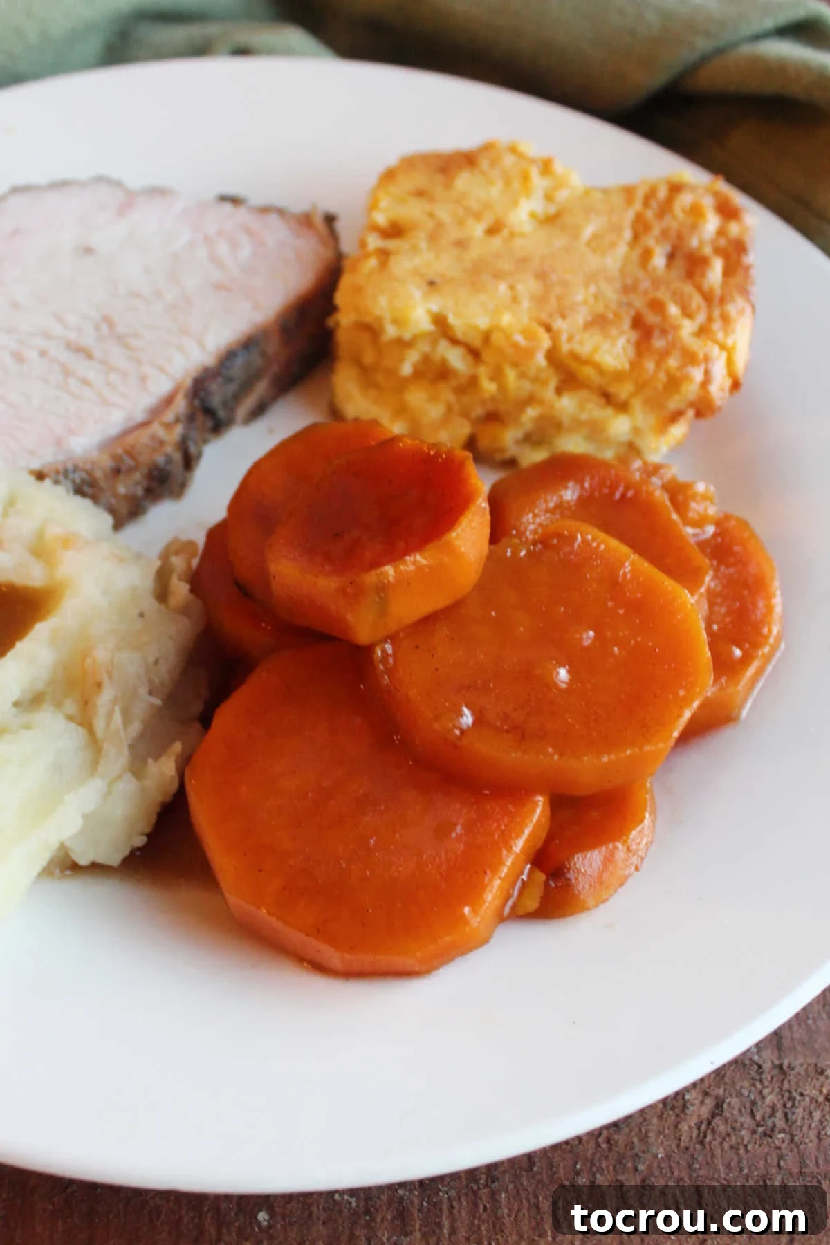 Close up of candied sweet potato slices on plate with corn casserole, smoked turkey breast, and mashed potatoes and gravy. 