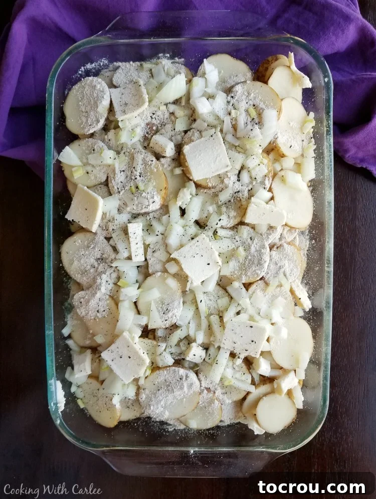 A clear glass casserole dish filled with layers of thinly sliced potatoes, patting of butter, and finely chopped onions, ready for baking.