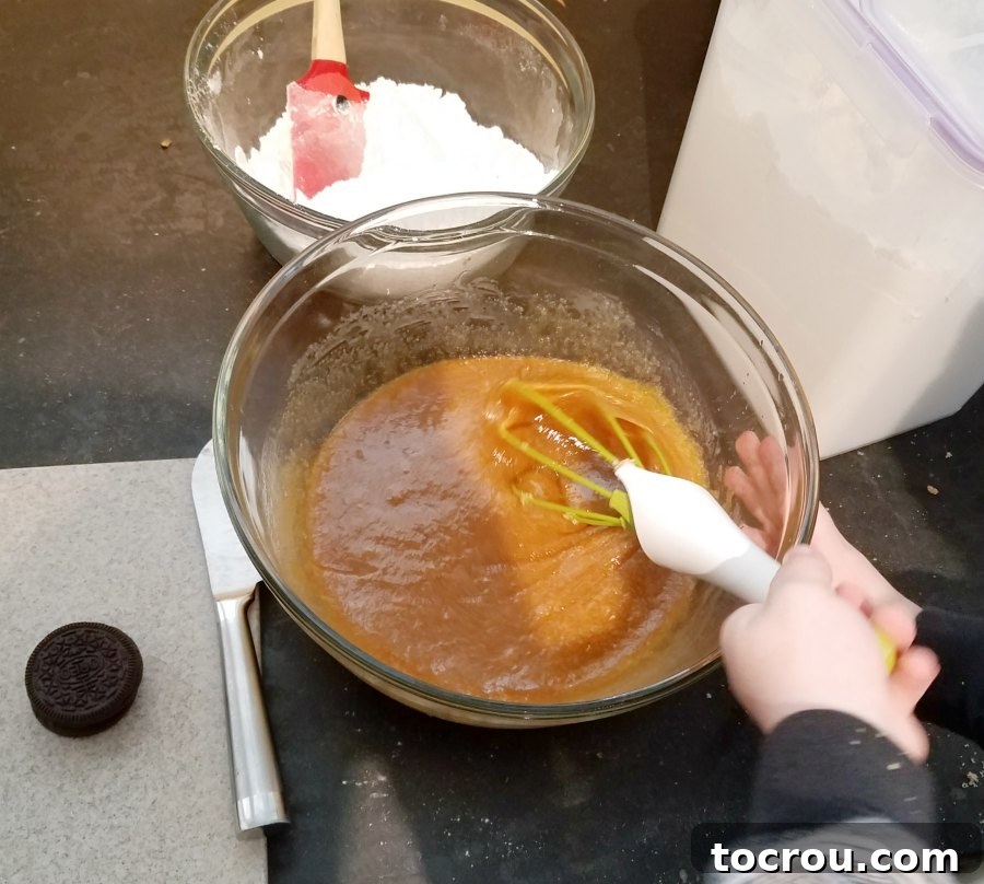 A child's hand actively stirring a bowl of blondie batter with a whisk, showcasing the smooth, creamy texture of the mixture.