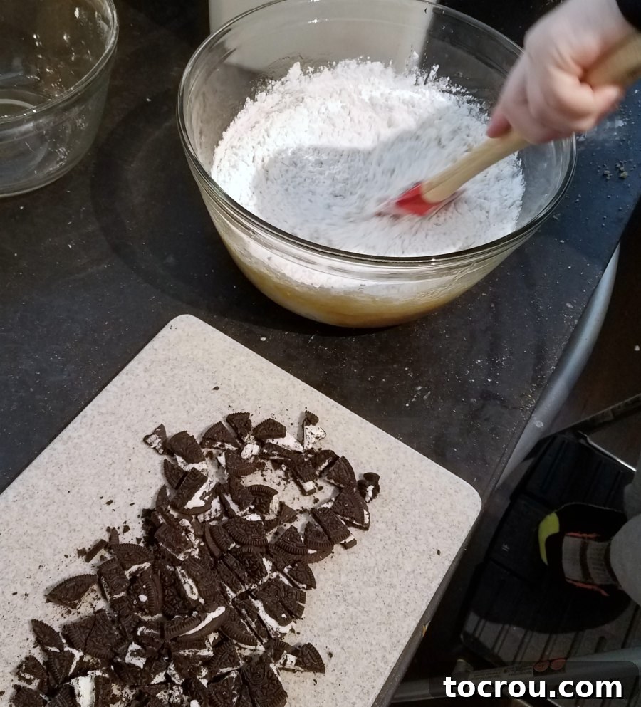 Smooth blondie batter being stirred with a spatula in a large bowl, with a cutting board holding roughly chopped Oreo cookies nearby.