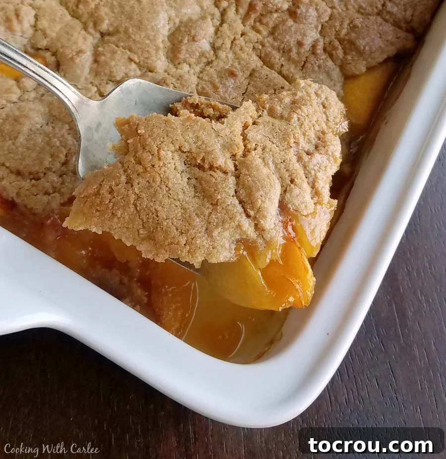 Freshly Baked Brown Sugar Peach Cobbler big serving spoon lifting out corner of freshly baked brown sugar peach cobbler.