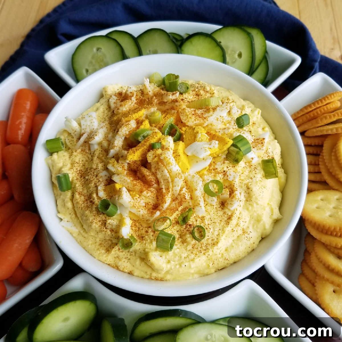 Bowl of deviled egg dip topped with paprika and green onion served with cucumber slices, baby carrots, and crackers.