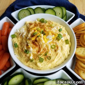 Bowl of deviled egg dip topped with paprika and green onion served with cucumber slices, baby carrots, and crackers.