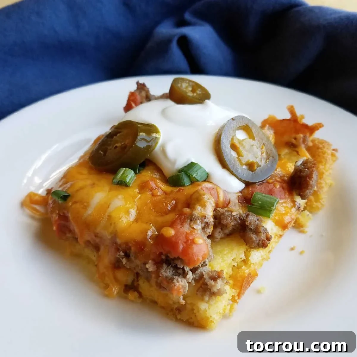 Delicious Homemade Turkey Tamale Casserole with Fresh Toppings A square portion of turkey tamale casserole, beautifully topped with fresh sour cream, vibrant sliced jalapenos, and finely chopped chives, presented on a white plate ready to eat.