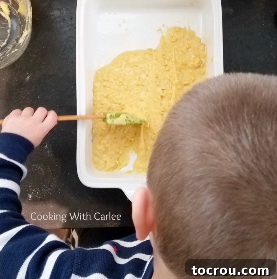 Little Hands Helping to Prepare the Tamale Casserole Base Child's small hands carefully spreading a light yellow cornmeal mixture into an oven-safe casserole dish, highlighting a hands-on cooking moment.