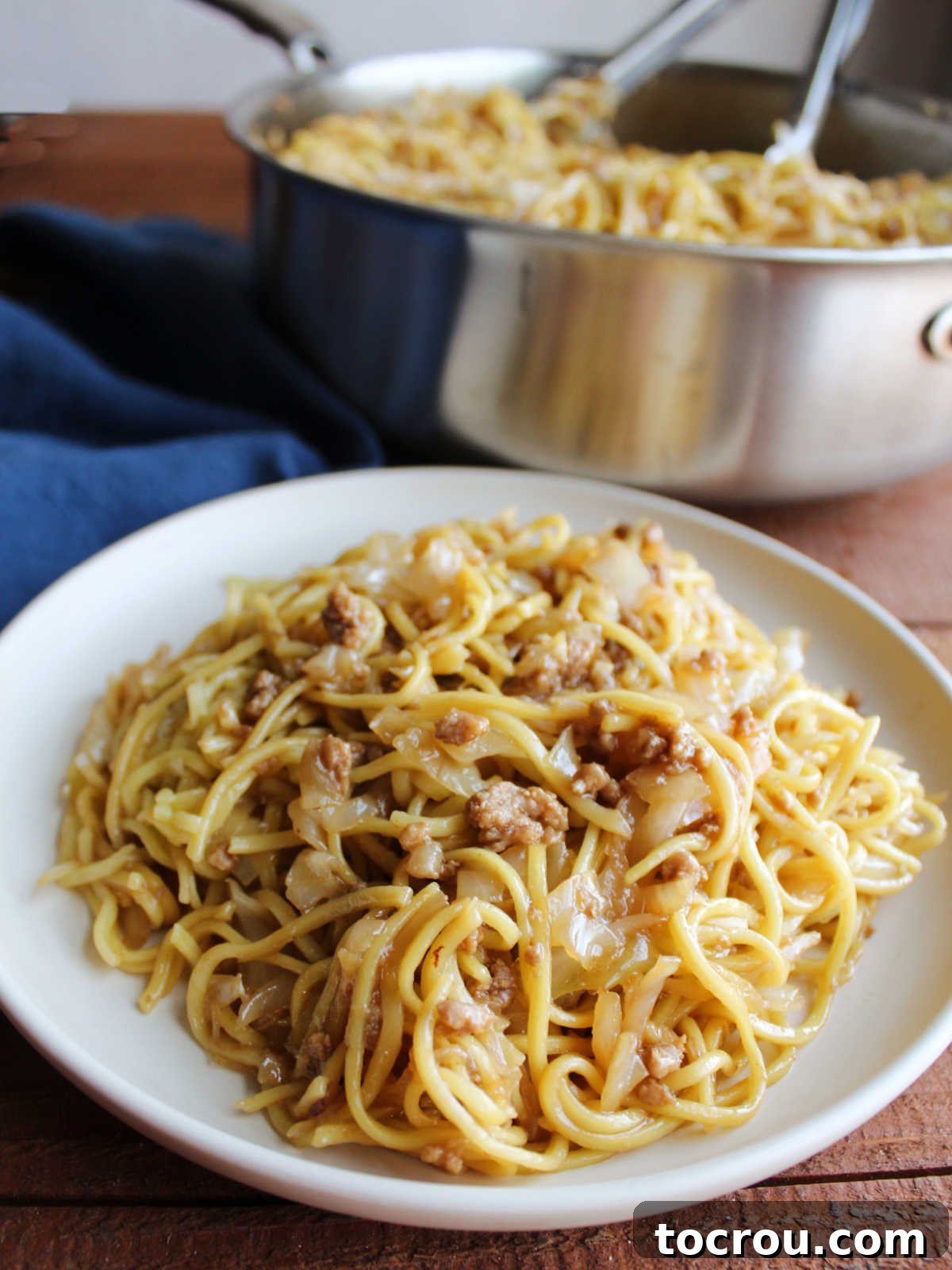 A close-up view of a plate piled high with glistening chow mein noodles, coated in a sticky sauce and mixed with tender ground pork, cabbage, and onions, highlighting the delicious texture.