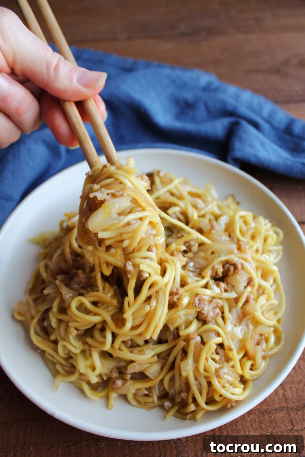 A hand expertly uses chopsticks to gather a generous portion of glossy chow mein noodles, perfectly cooked cabbage, and tender ground pork, ready to be eaten.