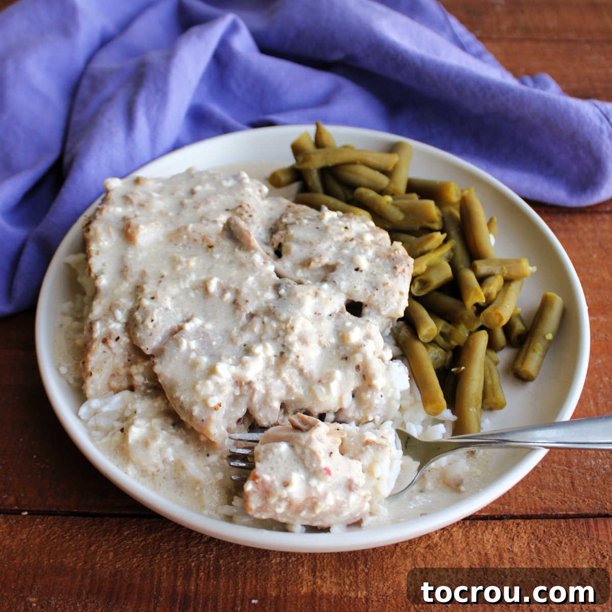 Fork going through tender pork chop in creamy ranch sauce served over rice with a side of green beans, highlighting the dish's irresistible texture.