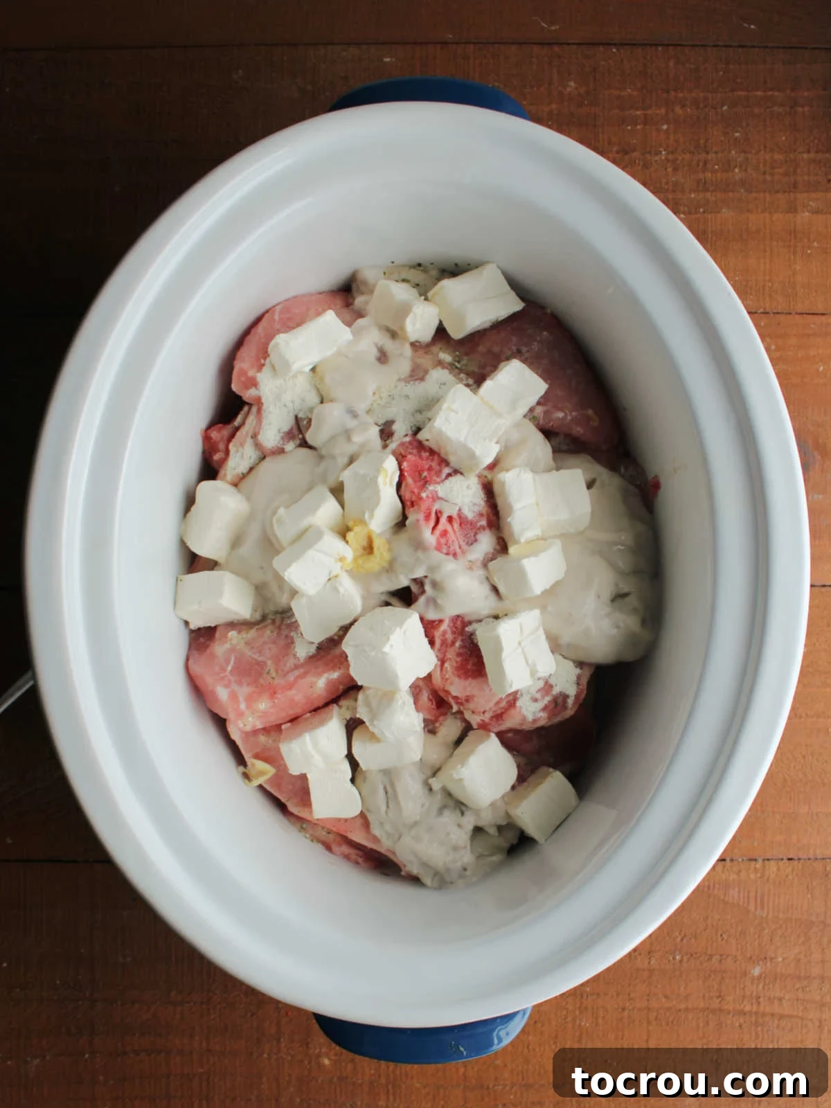 Crockpot with pork chops covered in ranch seasoning, cream of mushroom soup, and cream cheese, forming layers ready to be cooked slowly.