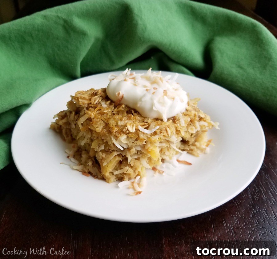 A square piece of pineapple and coconut baked oatmeal topped with a generous dollop of creamy yogurt and a sprinkle of golden toasted coconut.