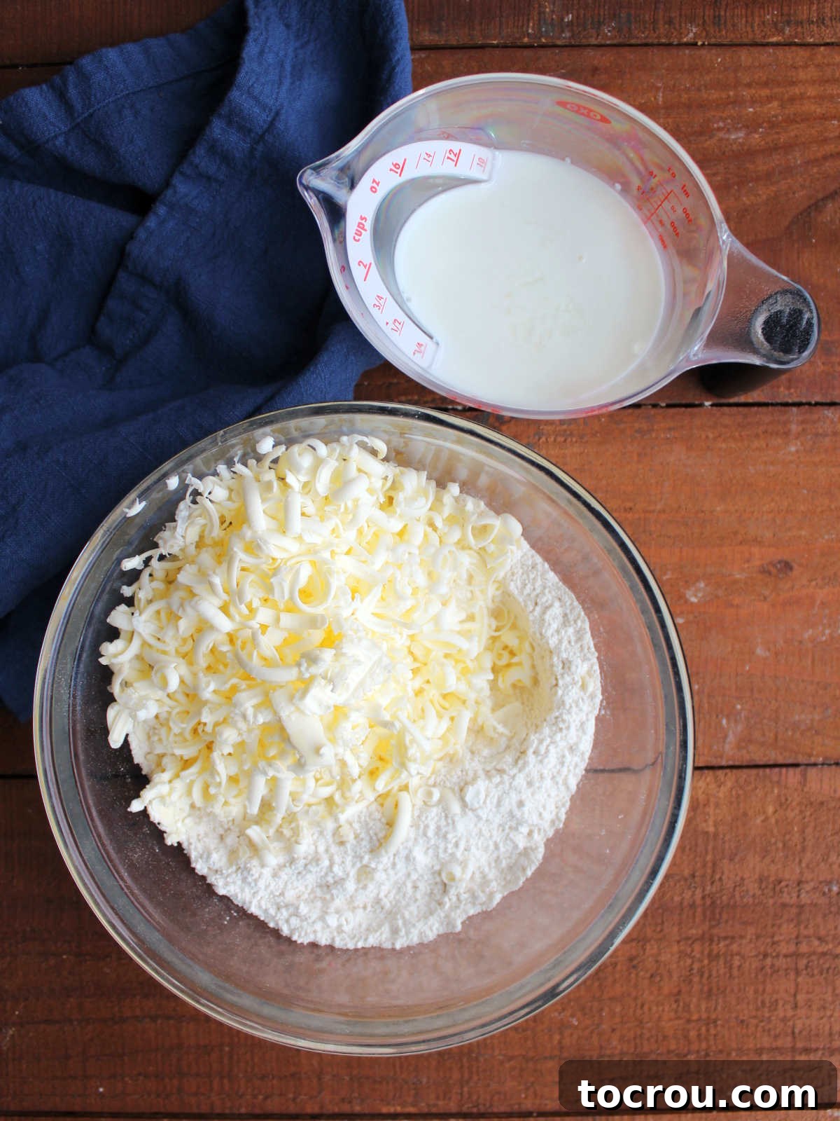 Bowl with flour, baking powder and salt mixture with grated frozen butter on top next to measuring cup with soured milk inside. Visualizing the initial ingredients for biscuit making.