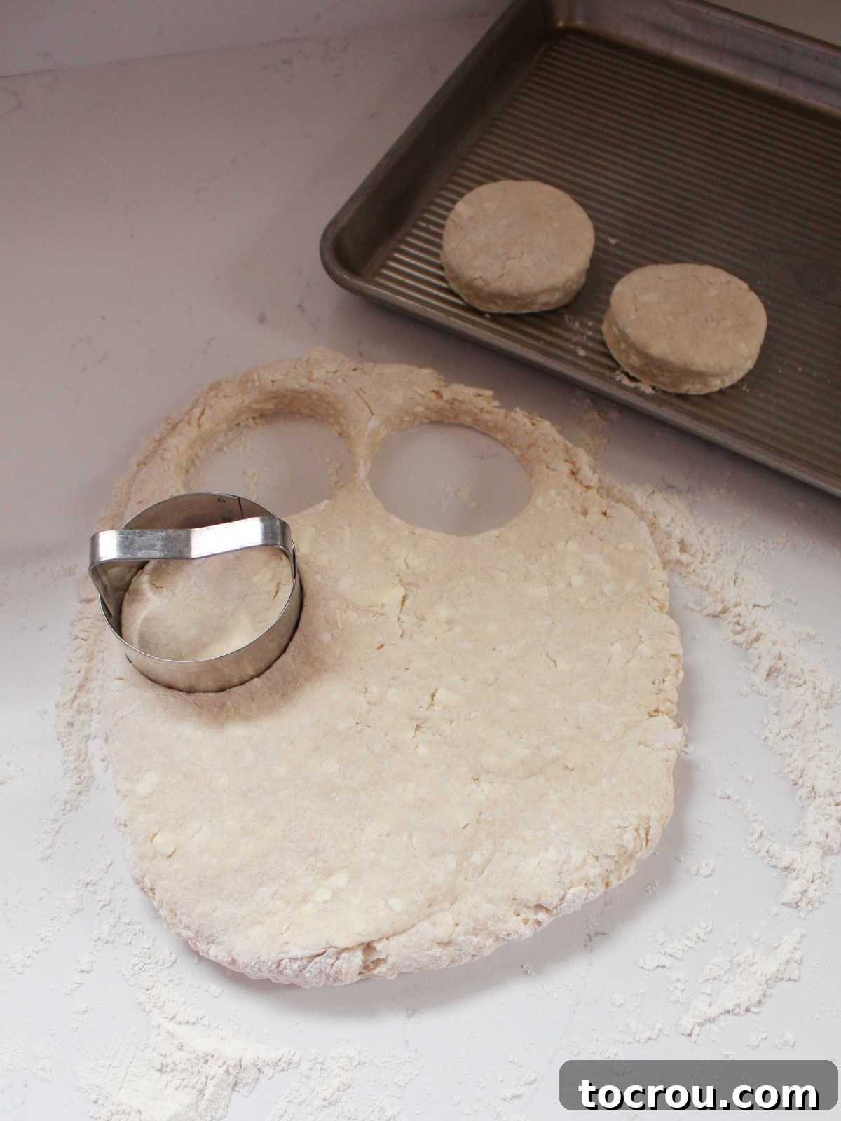 Biscuit cutter getting circles of dough out with some on a baking sheet nearby. Demonstrating the cutting technique.