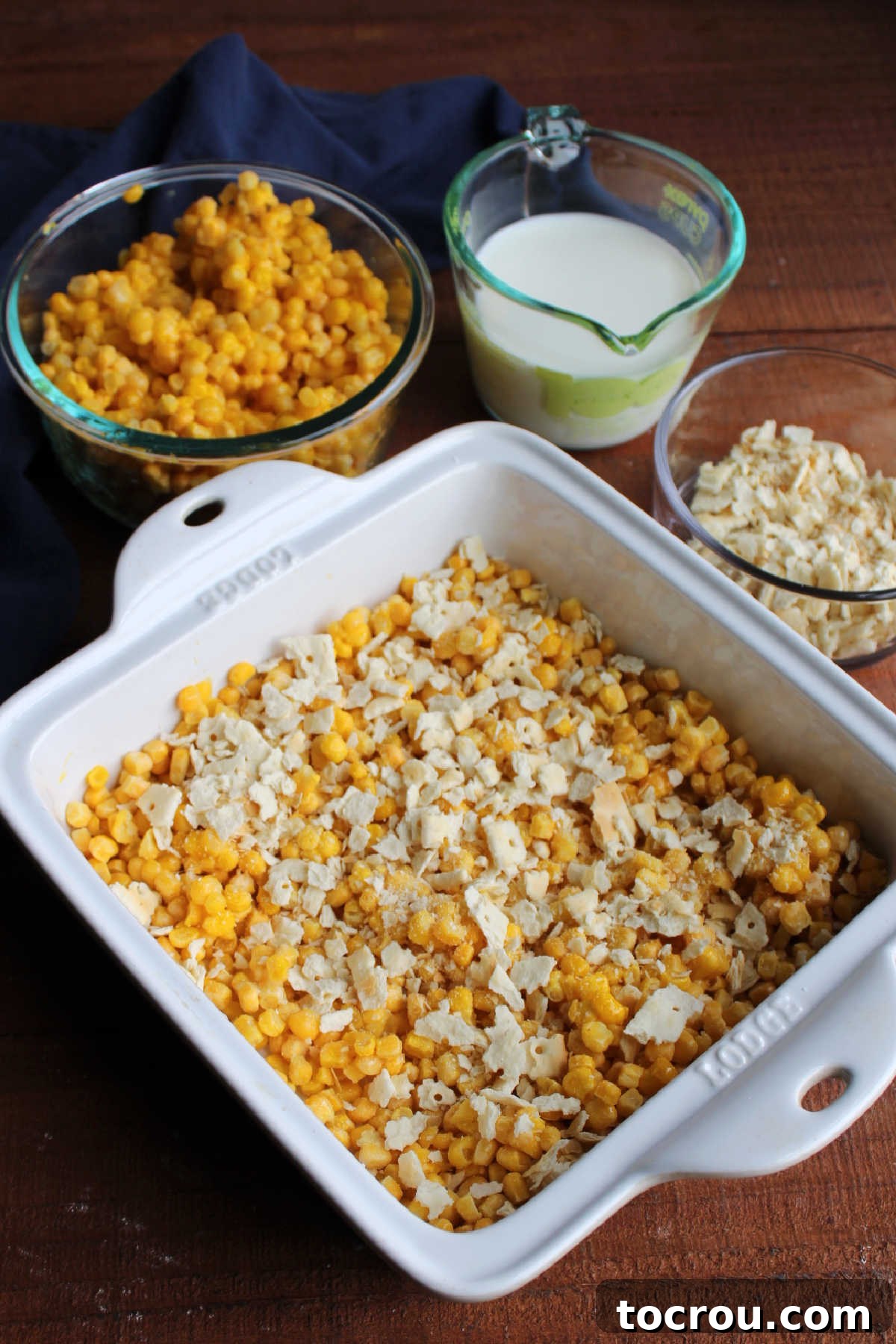 Assembling the casserole: Corn, eggs, and crackers in a baking dish. A square casserole dish filled with corn, beaten eggs, and cracker crumbs, with other ingredients visible in the background, ready for mixing.