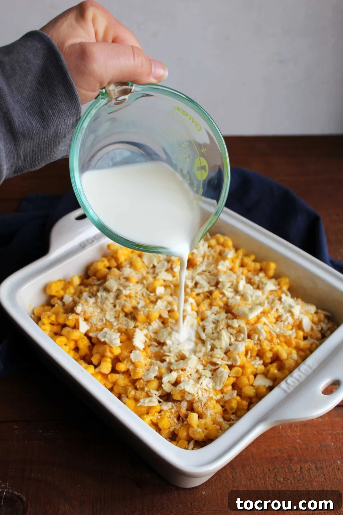 Finishing touch: Pouring milk over the scalloped corn casserole before baking. Hands pouring milk over the layered corn, egg, and cracker crumb mixture in a casserole dish, just before baking.