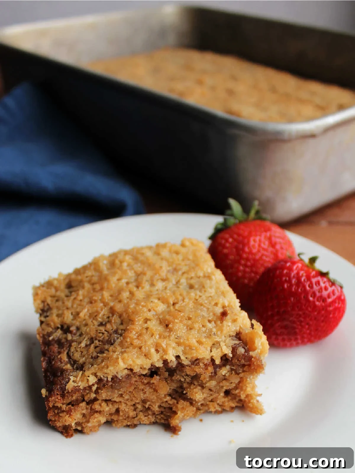 Piece of oatmeal cake with broiled coconut topping served on plate with strawberries with cake pan and remaining cake in the background. 