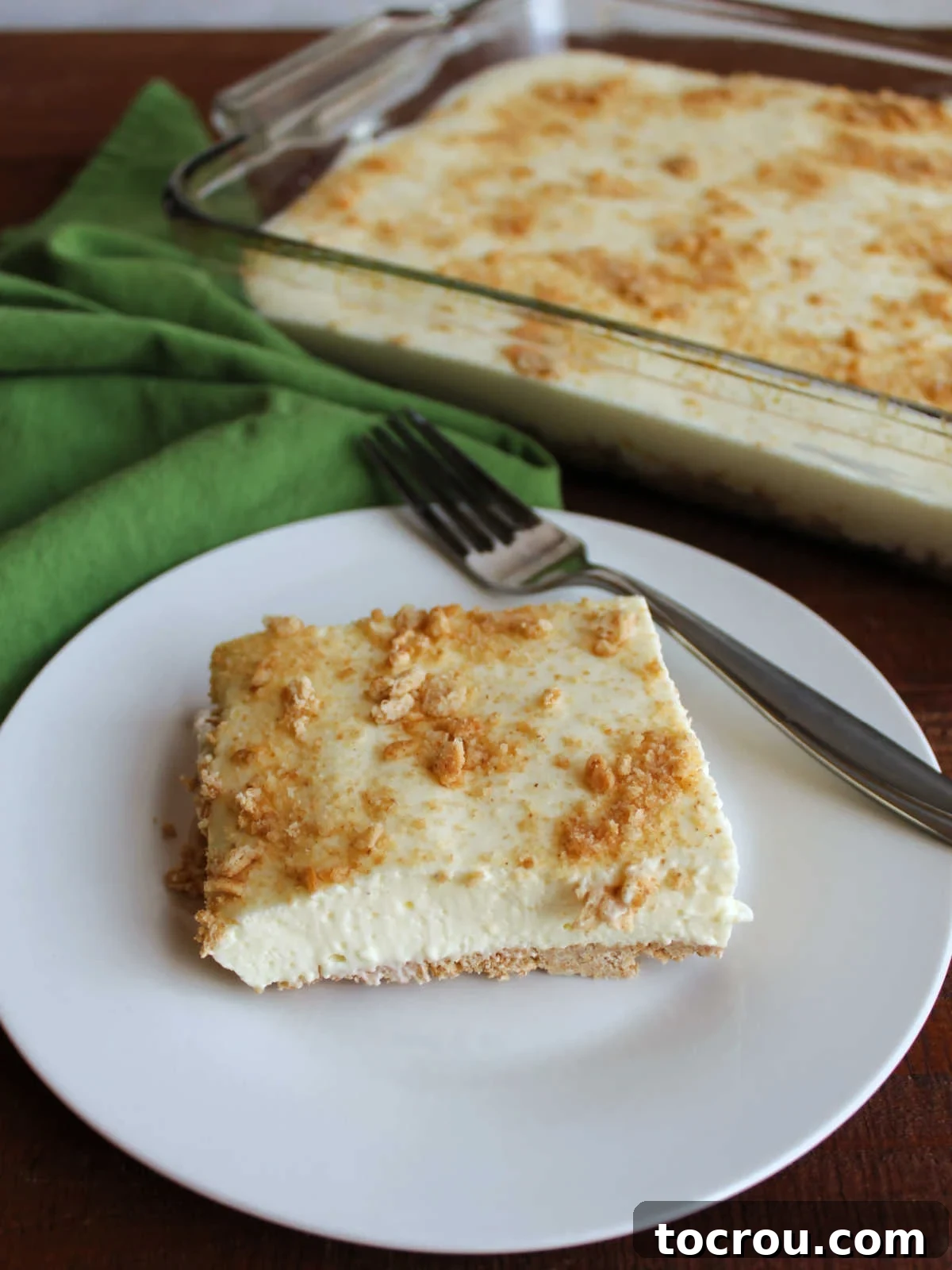 Slice of no bake cheesecake on plate with fork with 9x13-inch pan of remaining pale yellow cheesecake in the background.