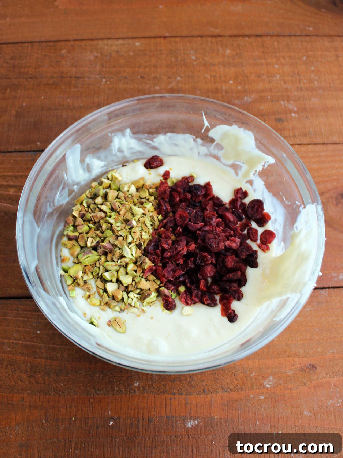 Glass mixing bowl with warm white fudge mixture after the chips have melted with the condensed milk topped with dried cranberries and pistachios, ready to be stirred in. 