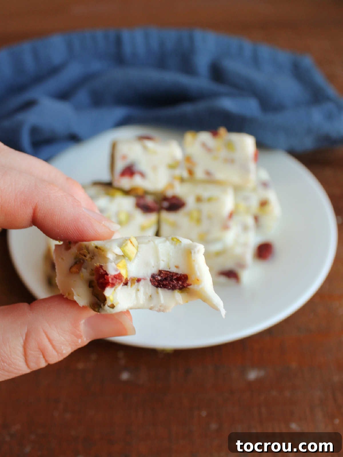 Hand holding piece of white fudge with a bite missing showing the creamy texture and cranberries and pistachios inside. 