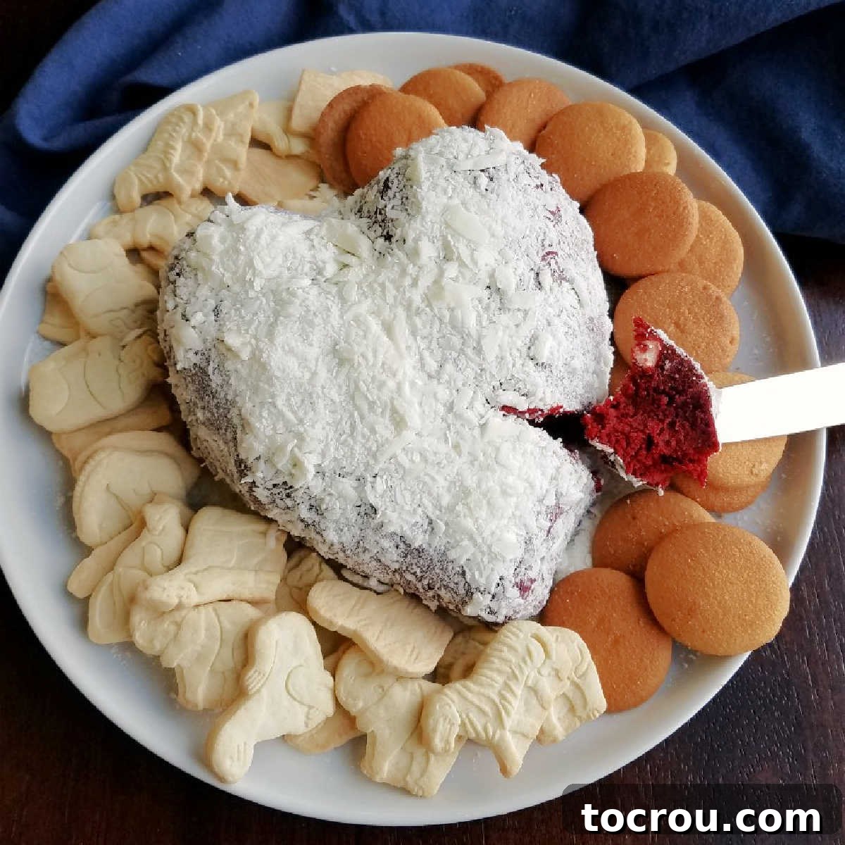 Luscious red velvet cake and cream cheese frosting flavors come together for a one of a kind dessert experience. Red velvet cheese balls are perfect for Valentine's Day, Christmas or just because! serving plate with heart shaped red velvet cheese ball coated in white chocolate shavings with vanilla wafers and animal crackers for eating.